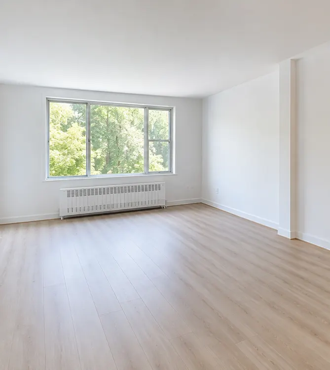 An empty, bright living room with pale wood-look floors, white walls and a wide window onto trees.