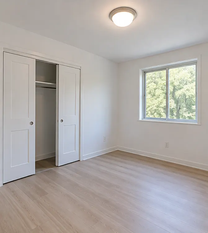 A bright empty bedroom with a sliding-door closet, a ceiling light and a window onto green trees.