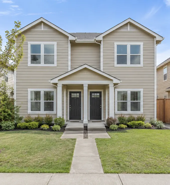 A tan two-story duplex with two black front doors under a shared porch and a small front lawn.
