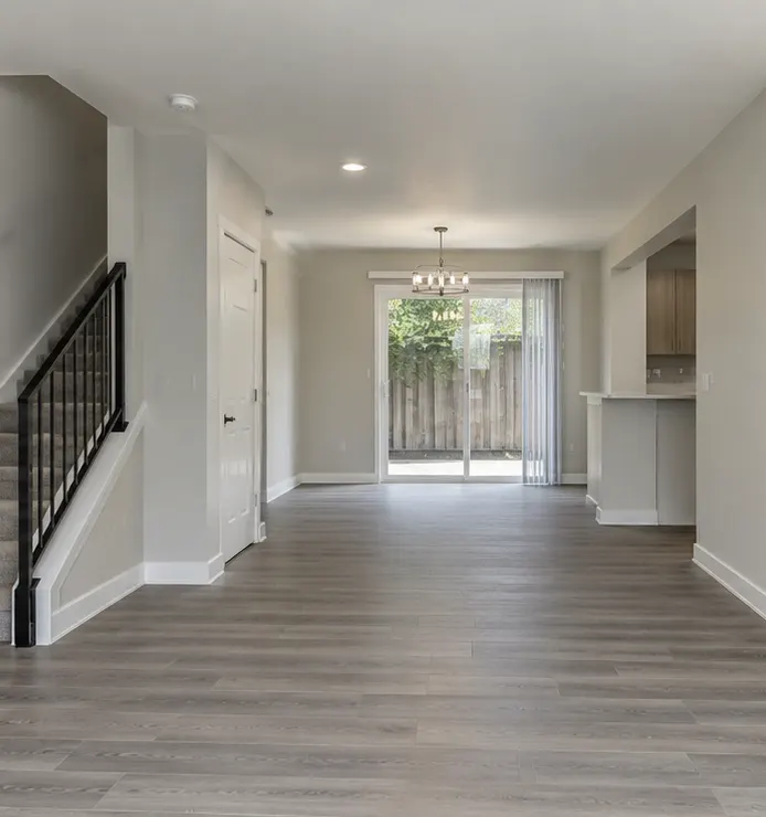 An empty townhouse living and dining room with gray plank floors, a stair rail and a sliding door to a patio.